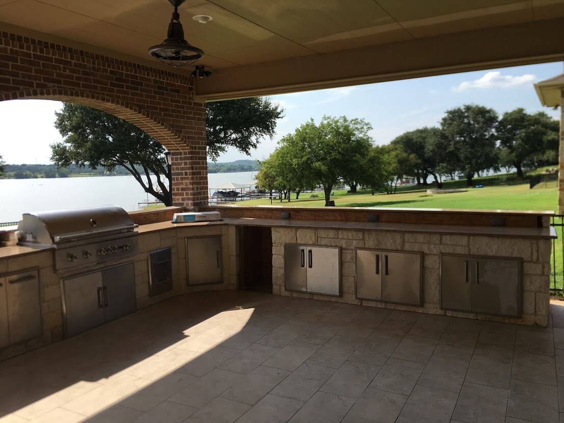 Outdoor kitchen with a built-in grill and stone cabinets under a covered patio, overlooking a lake