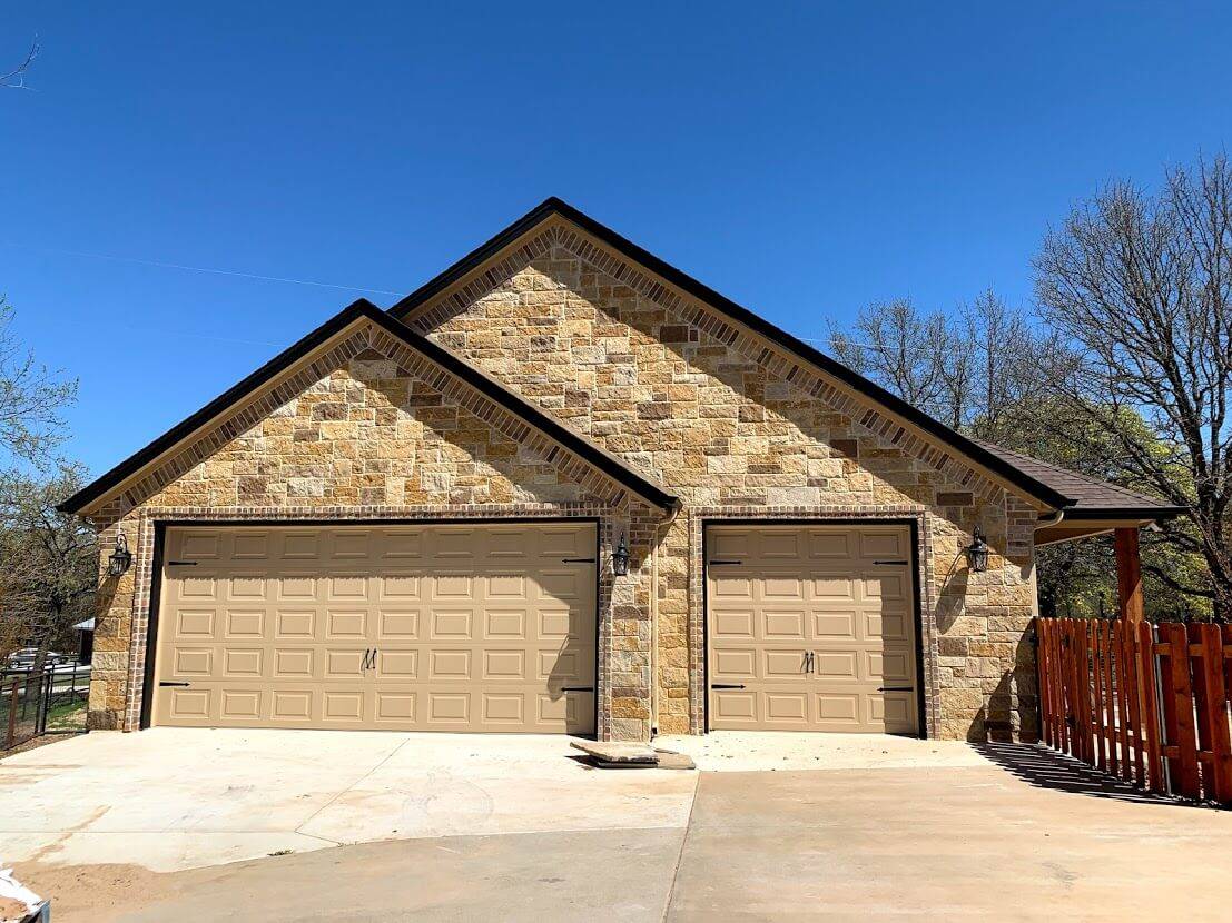 Stone garage with two gables, brick trim and two tan garage doors beside a cedar fence